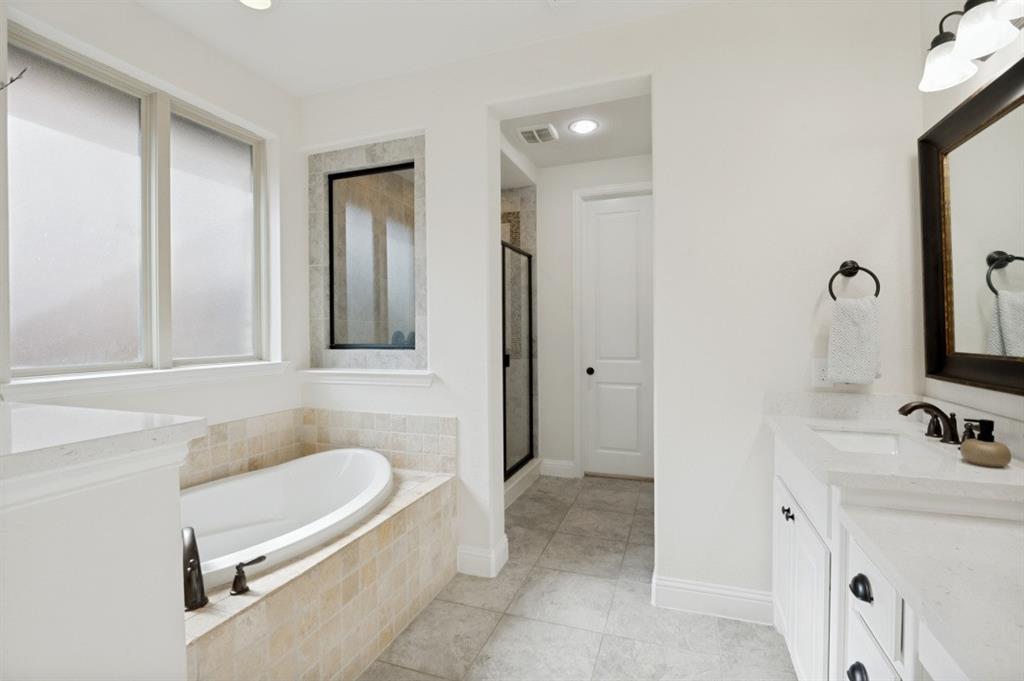 14215 Raspberry Lane Frisco, TX 75035 - Photo 11 of 30 Bathroom featuring a freestanding tub with tile surround, dual vanity with white countertops, framed mirror, and a glass-enclosed shower with black trim