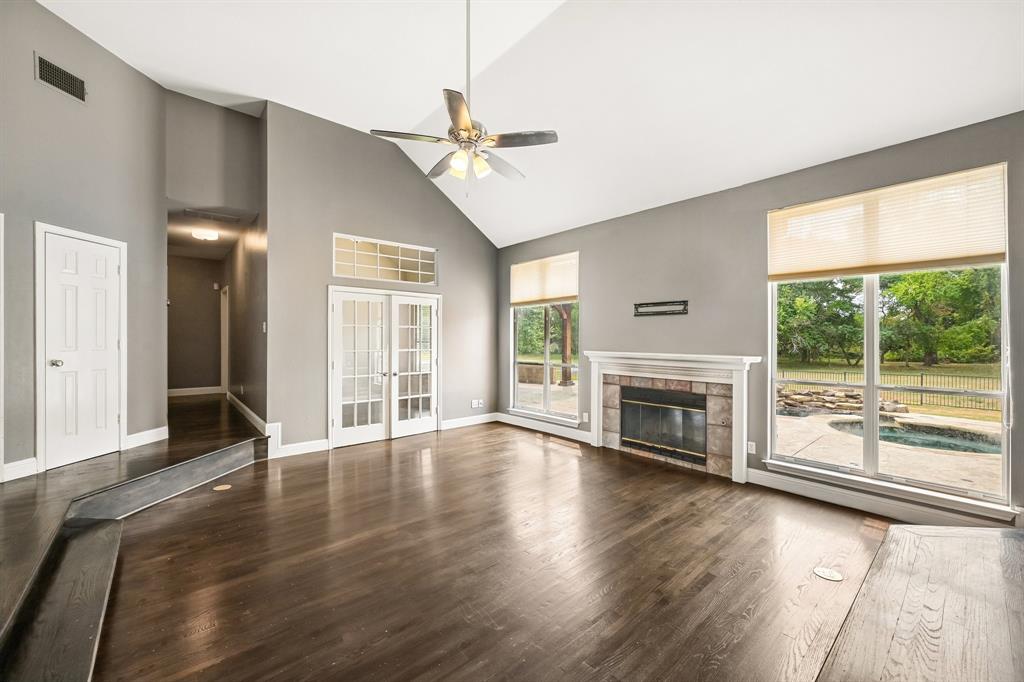 Vaulted ceiling with a ceiling fan, dark wood-finish flooring, stone-faced fireplace with a white mantel, and large windows revealing an outdoor pool and patio