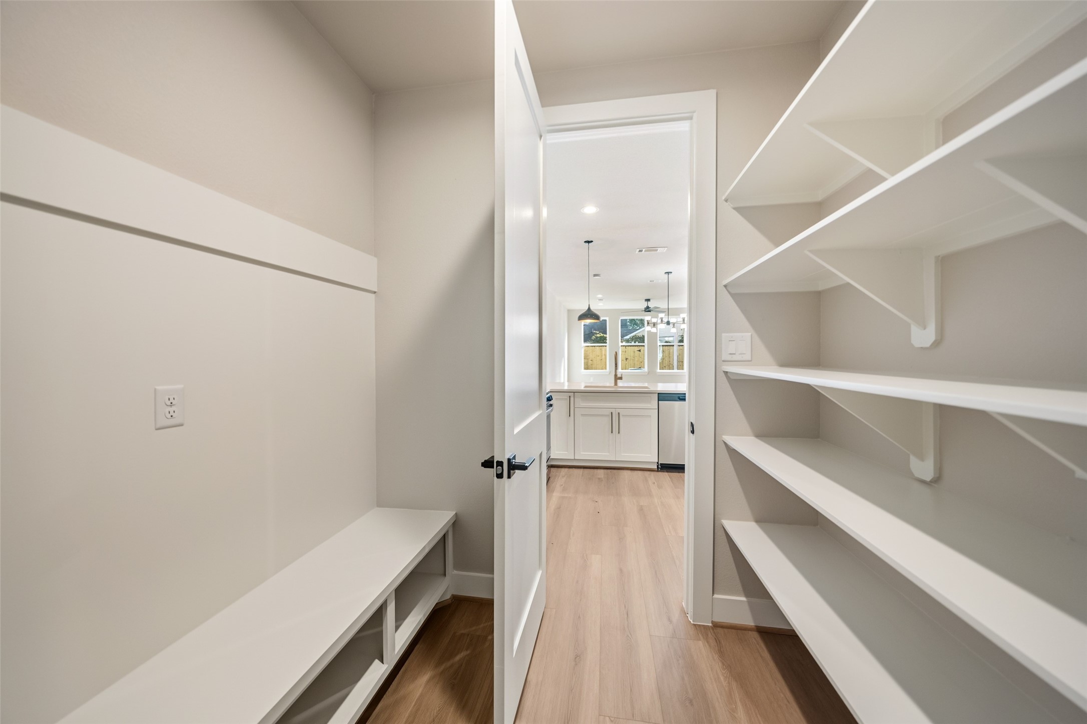 3422 Elser Street Houston, TX 77009 - Photo 10 of 26 This photo showcases a bright pantry or storage area with white shelving on one side and a bench with storage underneath on the other. The space leads into a kitchen with modern pendant lighting and a view of large windows, providing a spacious and open feel.