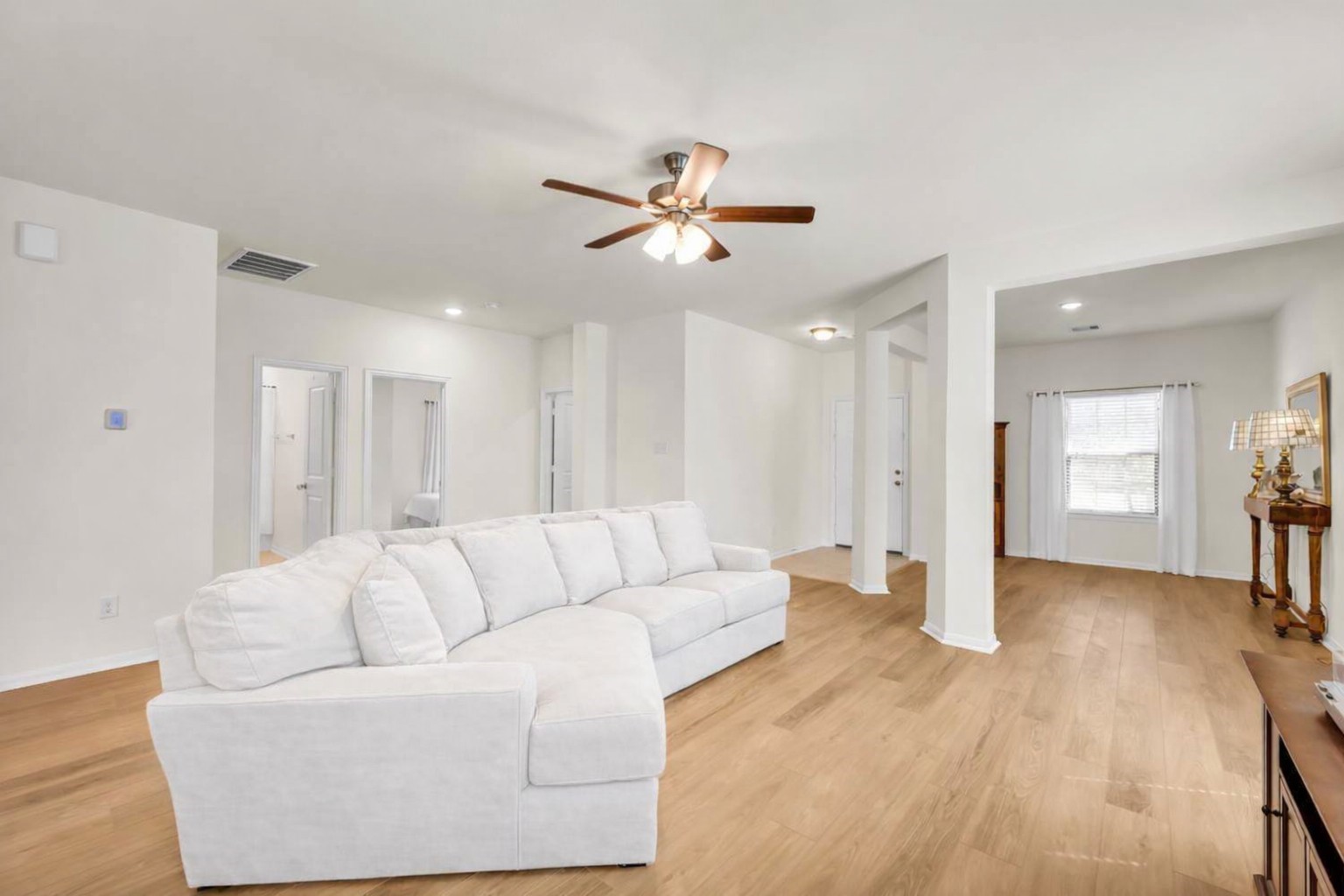 This spacious living area features new flooring, neutral walls, and ample natural light. It includes a large white sectional sofa and ceiling fan, providing a comfortable and inviting space.