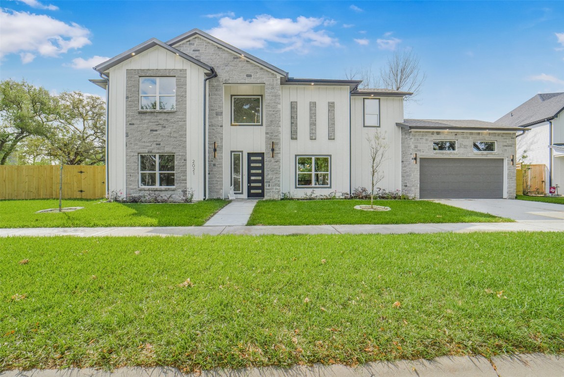 This modern two-story home features a sleek exterior with a blend of stone and siding, large windows, and a two-car garage. It's set on a well-maintained lawn with young trees and a paved walkway leading to the front door.