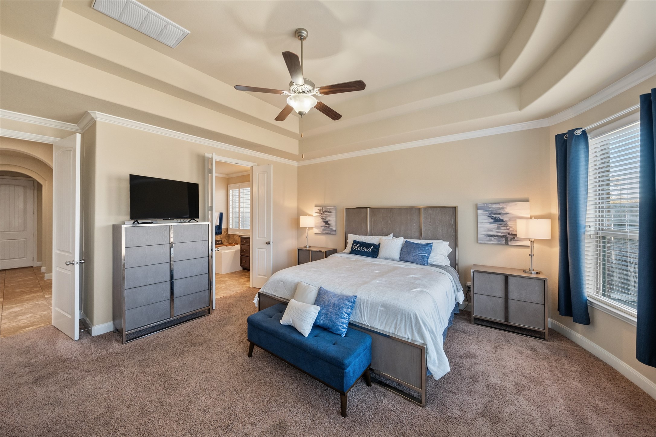 4747 Abercorn Street Rosharon, TX 77583 - Photo 20 of 46 This spacious primary bedroom features a tray ceiling with a ceiling fan, large windows for natural light, and plush carpeting. It includes a modern dresser, a cozy seating area at the foot of the bed, and an ensuite bathroom, providing comfort and style.