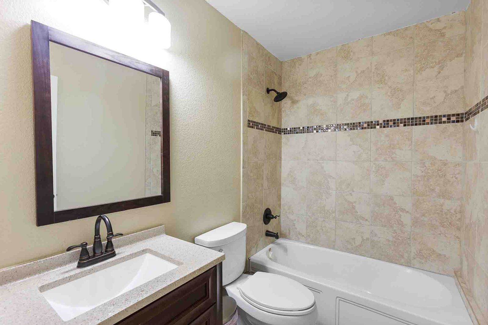 323 Woodbrook Trail Buda, TX 78610 - Photo 16 of 40 Bathroom featuring a white porcelain tub, tiled shower surround with decorative mosaic accent, dark wood-finish vanity with an integrated rectangular sink, light-colored speckled countertop, and bronze-finish fixtures