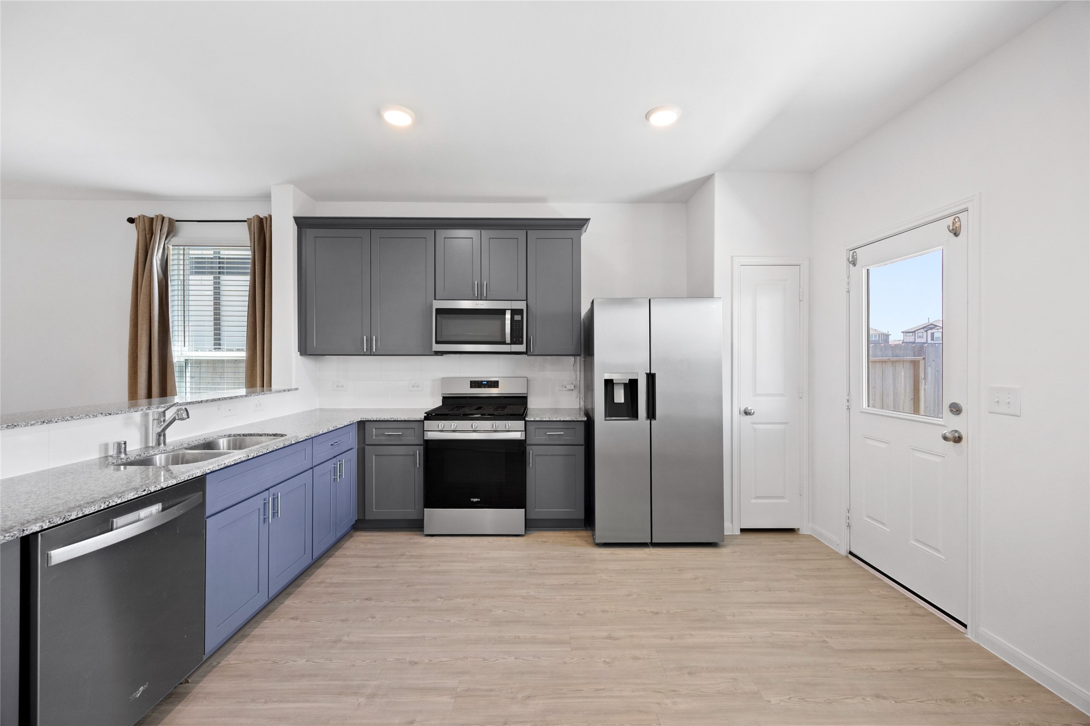 17907 Grain Auger Road Hockley, TX 77447 - Photo 2 of 24 Modern kitchen with sleek gray and blue cabinetry, stainless steel appliances, and a dual sink. Features a bright, open layout with wood-look flooring and a door leading outside.