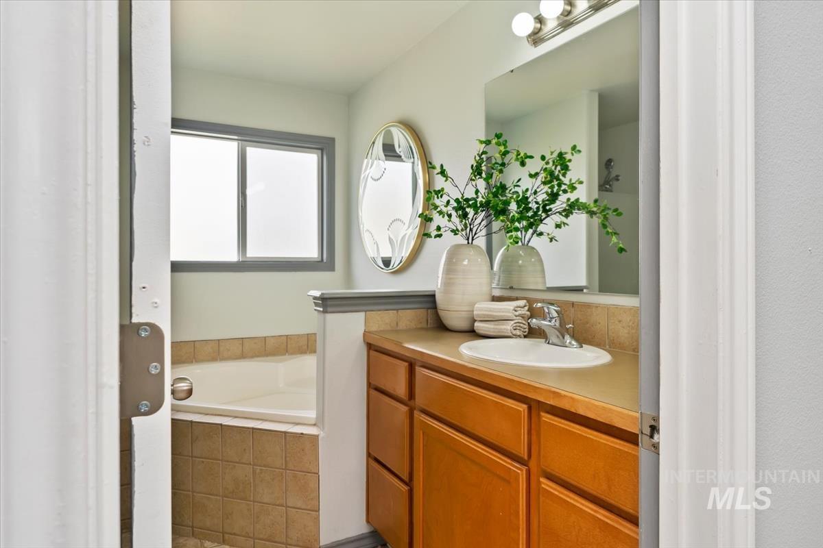 80 Cobblestone Court Middleton, ID 83644 - Photo 21 of 48 Bathroom featuring a corner soaking tub with tile surround, a frosted window, a vanity with wood-finish cabinetry, a single basin sink, and a large mirror