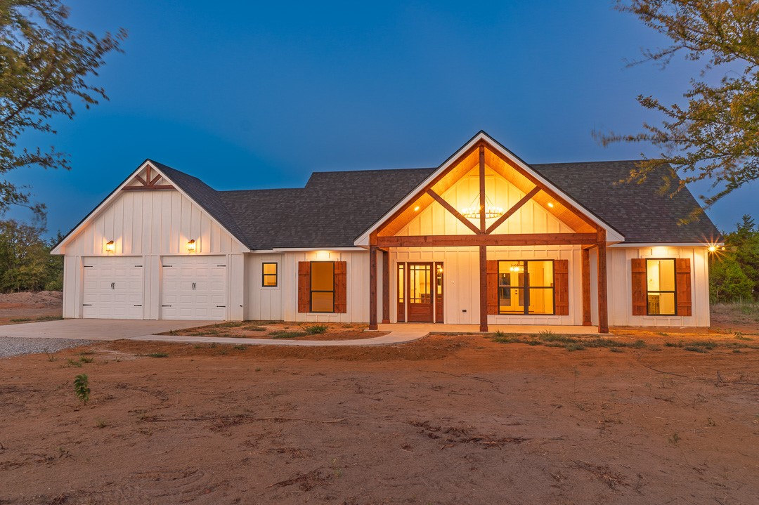 205 Blessings Way Colbert, OK 74733 - Photo 1 of 24 White board-and-batten siding with dark trim, dual garage doors, a covered entry with exposed wood beams, and decorative window shutters