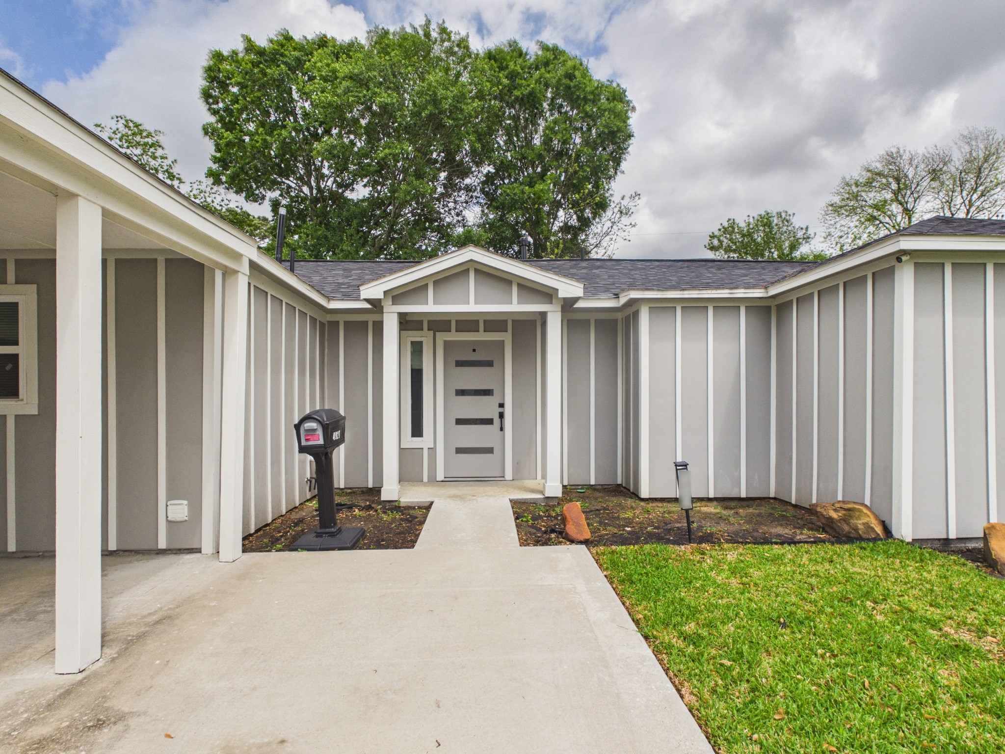 4927 Kelso Street Houston, TX 77021 - Photo 6 of 45 This photo showcases a modern, single-story home with a gray and white exterior, featuring vertical siding and a welcoming front entrance with a stylish door. The property includes a driveway, a neat lawn, and mature trees in the background, creating a charming curb appeal.