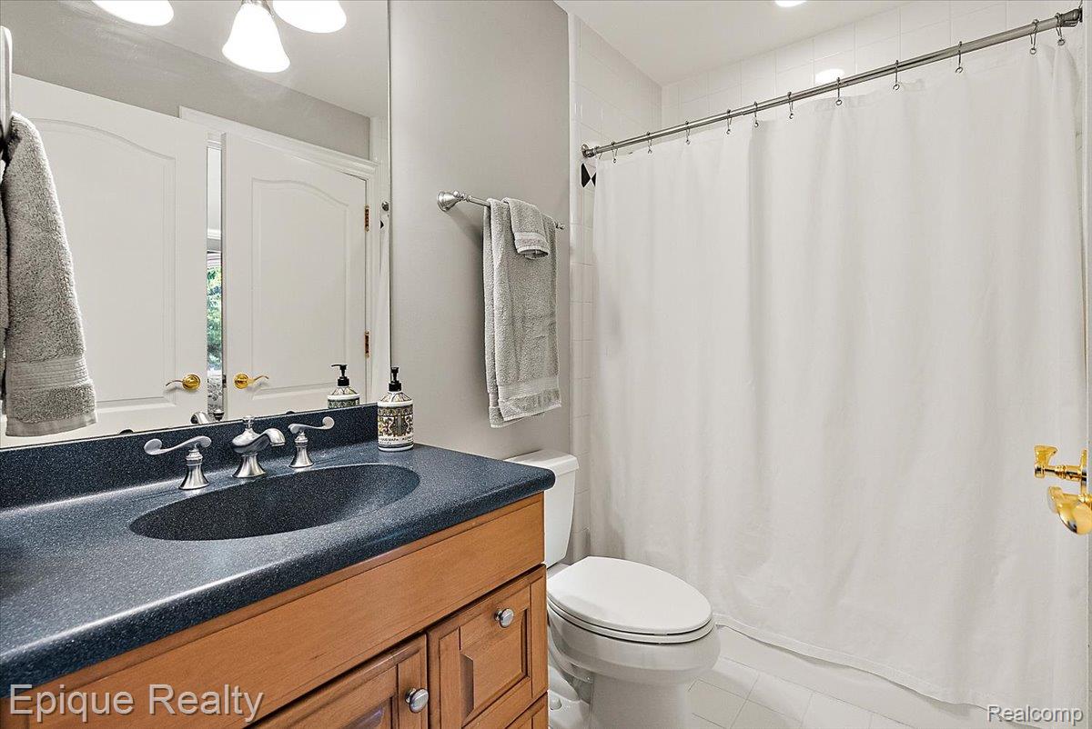 7662 Devins Ridge Clarkston, MI 48348 - Photo 65 of 81 Bathroom featuring a dark speckled countertop with an integrated sink, wood-finish vanity cabinetry, a shower with white tiled walls, and a white toilet