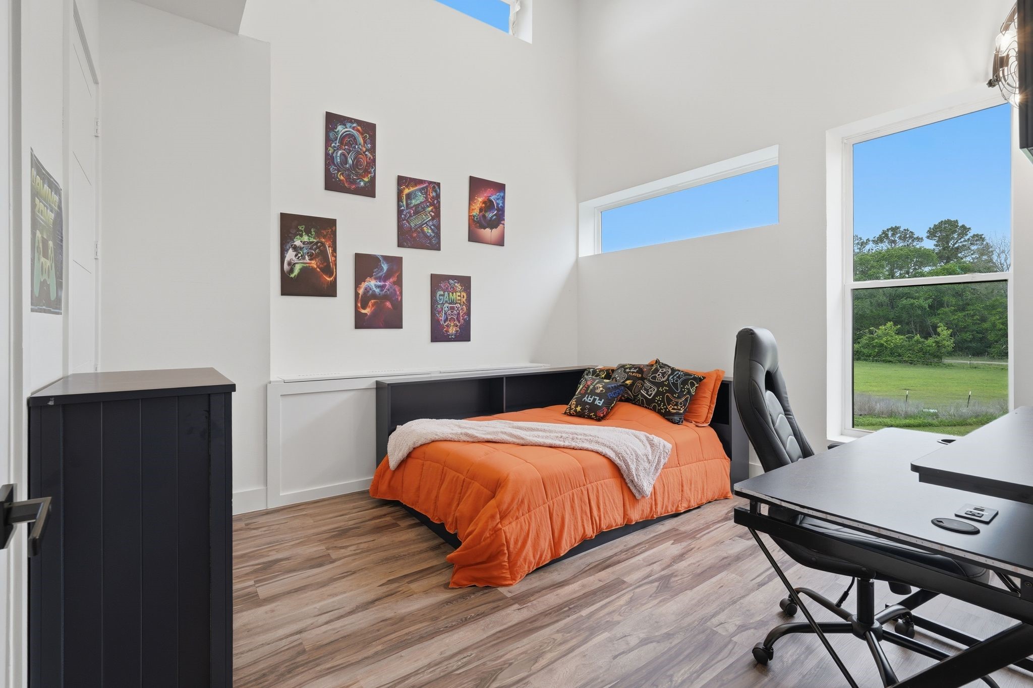2743 Bobville Road Montgomery, TX 77316 - Photo 39 of 50 Bright, modern bedroom with high ceilings and large windows offering scenic views. Features stylish wall art, a comfortable bed with orange bedding, and a sleek desk setup, ideal for work or study.