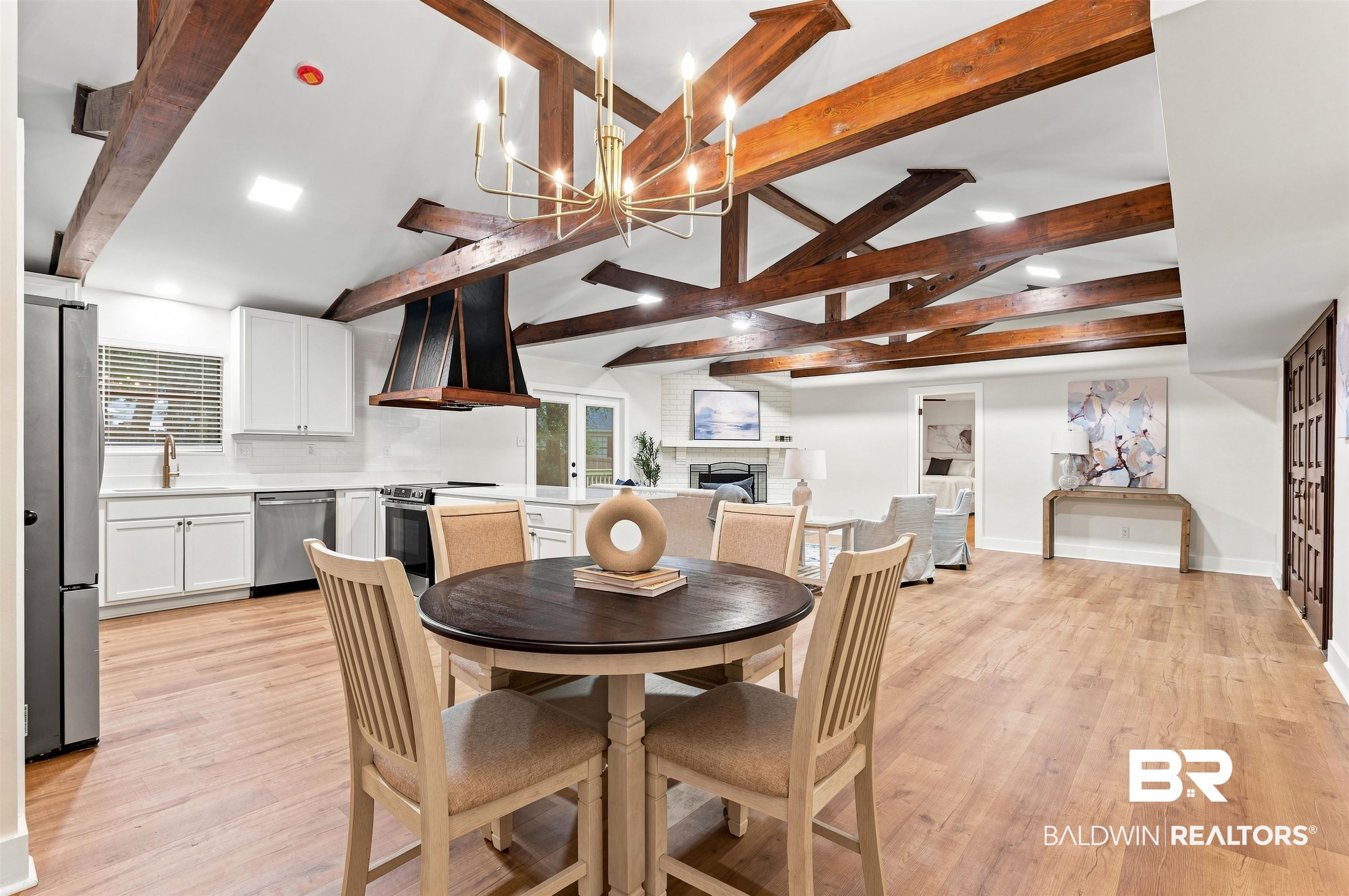 599 Ridgewood Drive Daphne, AL 36526 - Photo 8 of 41 Open-concept living area featuring exposed wood beams, wood-finish flooring, a contemporary chandelier, a kitchen with white cabinetry and stainless steel appliances, and a fireplace with a white mantel