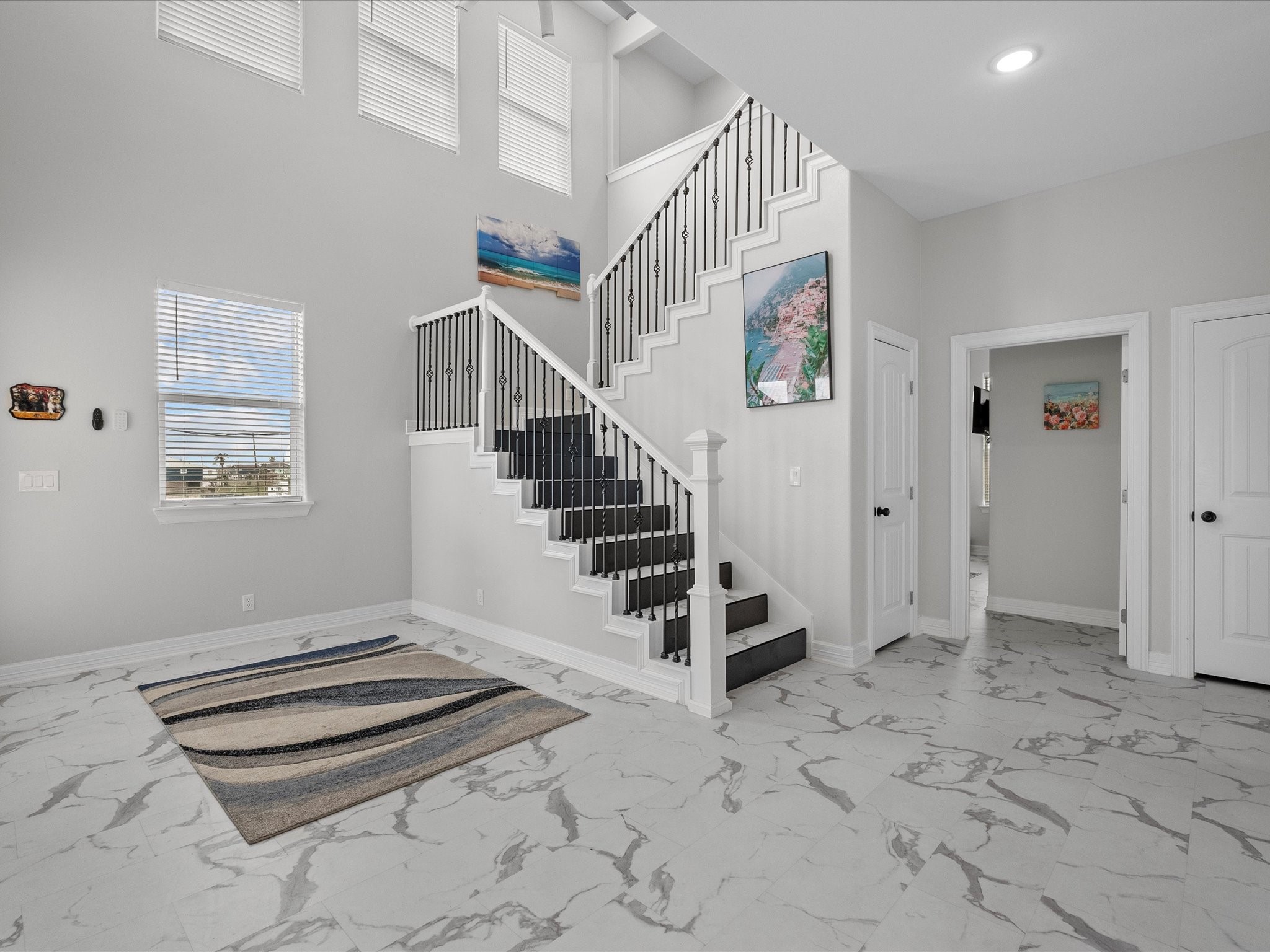 1018 Seashell Drive Surfside Beach, TX 77541 - Photo 12 of 49 Bright and spacious entryway featuring high ceilings, elegant staircase with black railing, and large windows. The marble-like tile flooring adds a modern touch, complemented by tasteful wall art, creating an inviting atmosphere.
