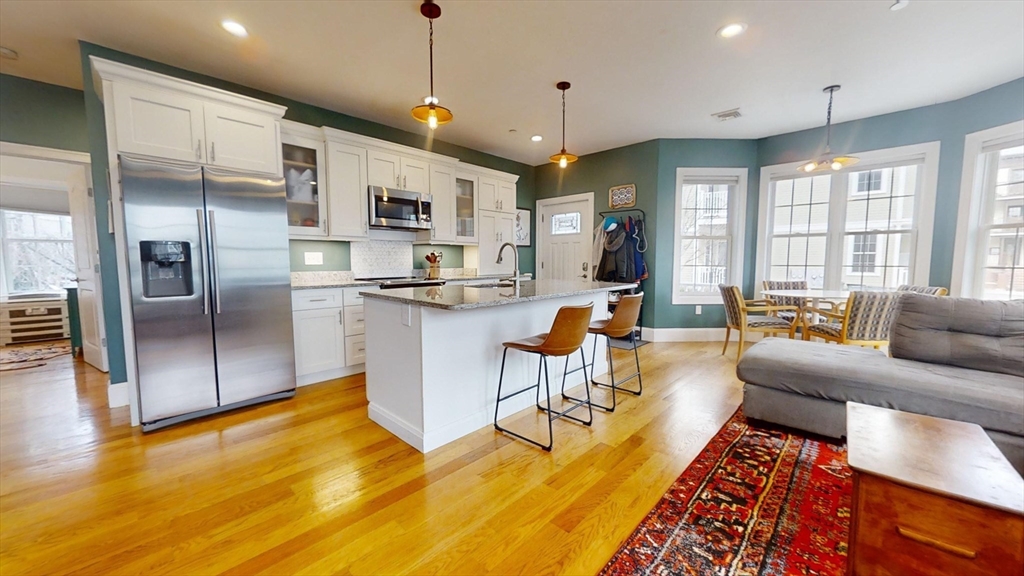 a living room with stainless steel appliances granite countertop a refrigerator a kitchen island a couches and a dining table with wooden floor