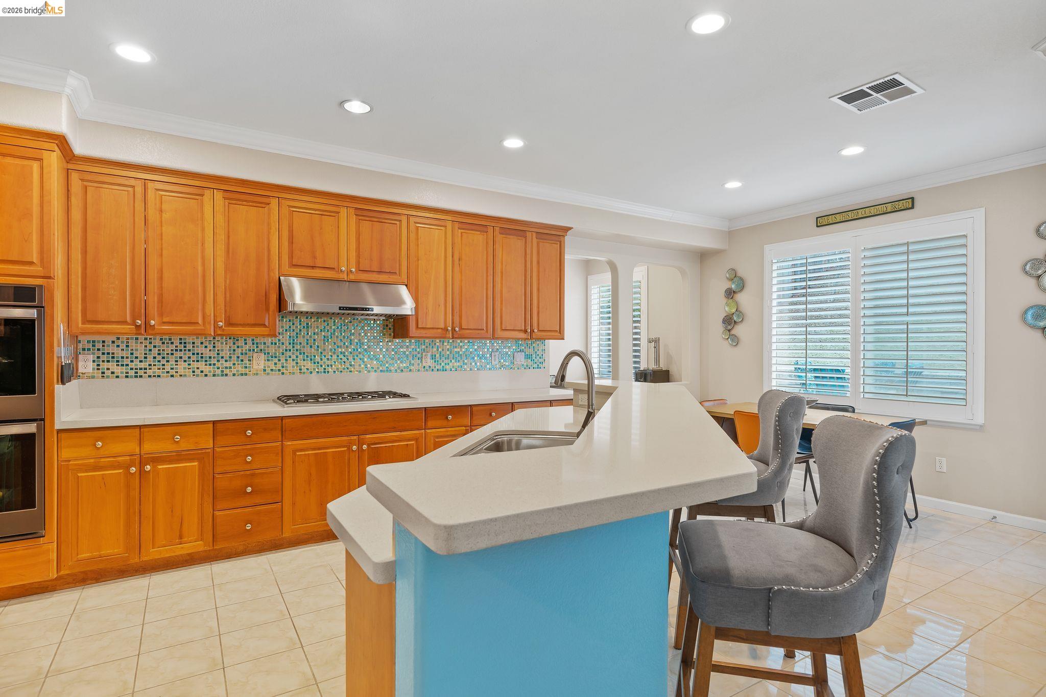 3237 Oso Grande Way Antioch, CA 94531 - Photo 12 of 47 Kitchen with wood finish cabinets, a kitchen island with sink, crown molding, light tile patterned flooring, and tasteful backsplash