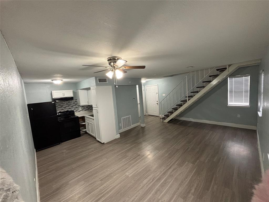 Open-concept living area featuring wood-finish flooring, a kitchen with white cabinetry and black appliances, a ceiling fan, and a staircase with metal railings