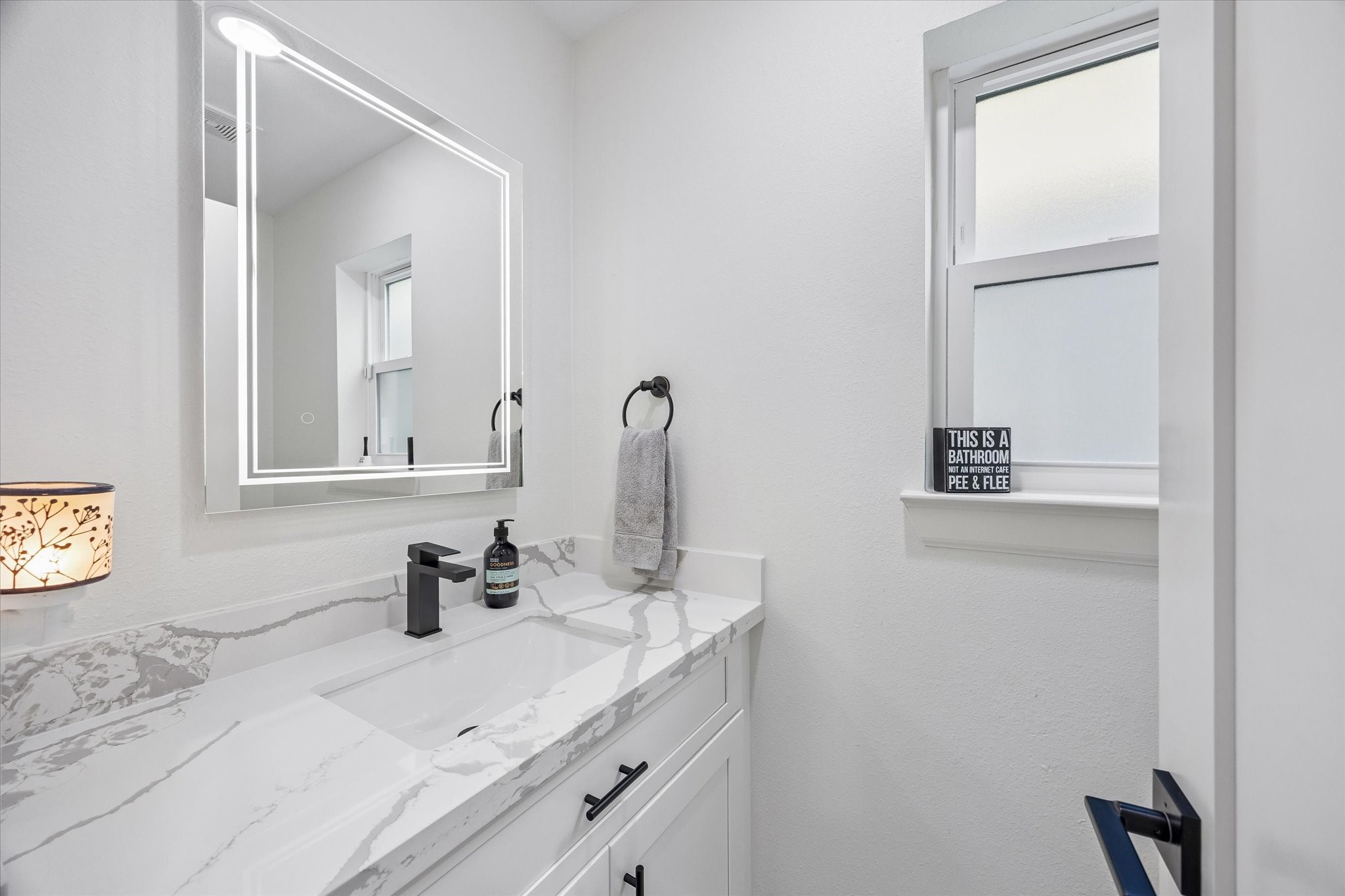 1420 Indiana Street Houston, TX 77006 - Photo 23 of 31 This is a modern bathroom featuring a sleek white vanity with a marble countertop, an illuminated mirror, and a frosted window for privacy. Simple and elegant design.