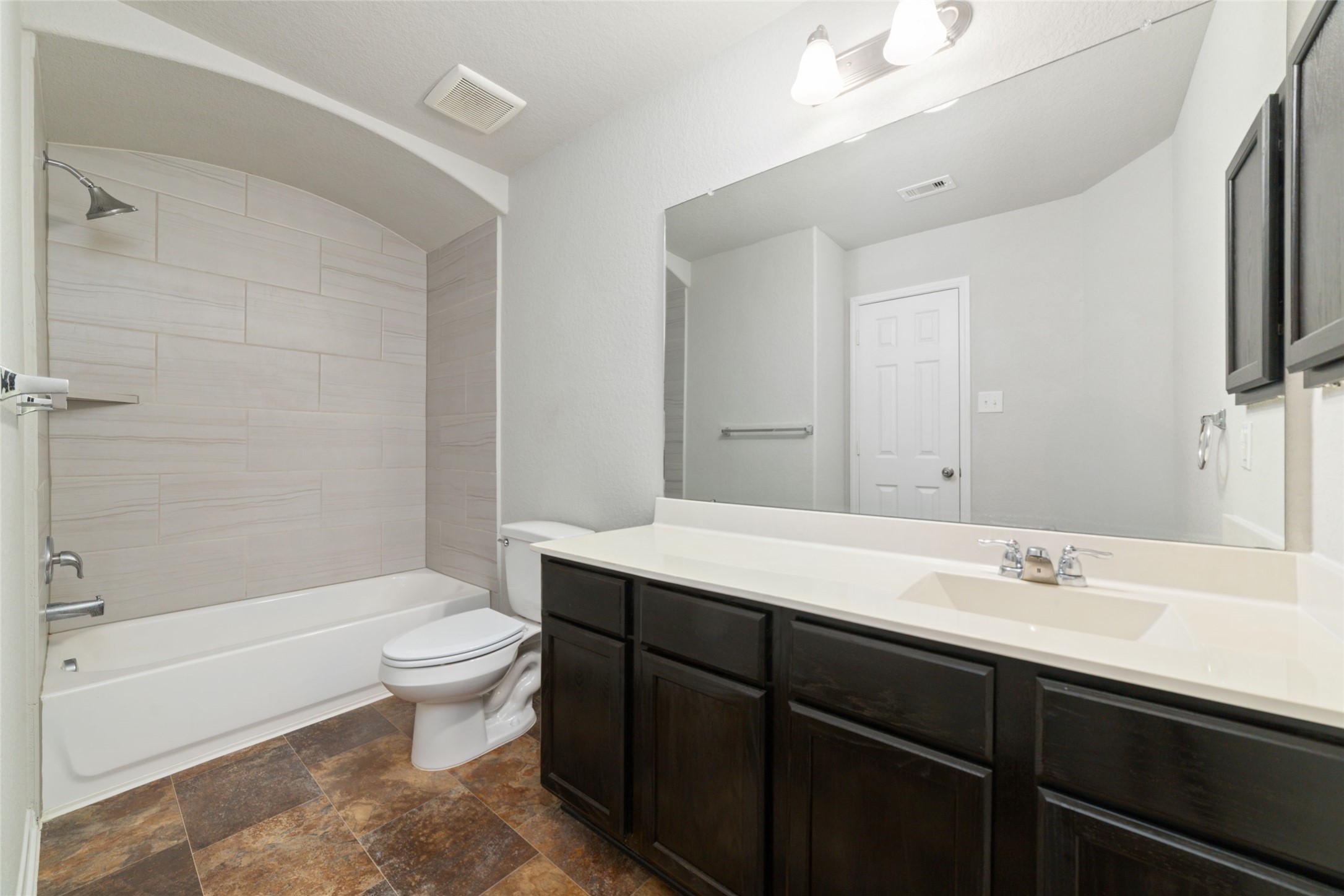 14838 Ruddy Duck Lane Houston, TX 77049 - Photo 25 of 33 This bathroom features a modern design with a large mirror, double sink vanity, and dark wood cabinets. It includes a bathtub with a tiled shower area and has a clean, neutral color palette. The floor is covered with patterned tiles, adding a touch of warmth.