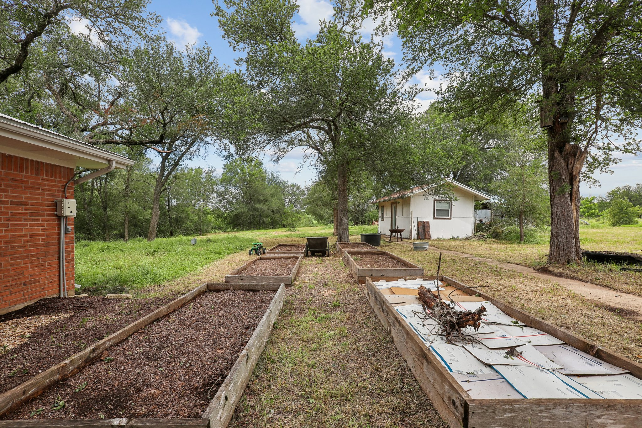 406 Monkey Road Elgin, TX 78621 - Photo 27 of 39 Expansive outdoor space featuring multiple raised garden beds, mature trees, a detached auxiliary building with white siding, and a brick-sided structure