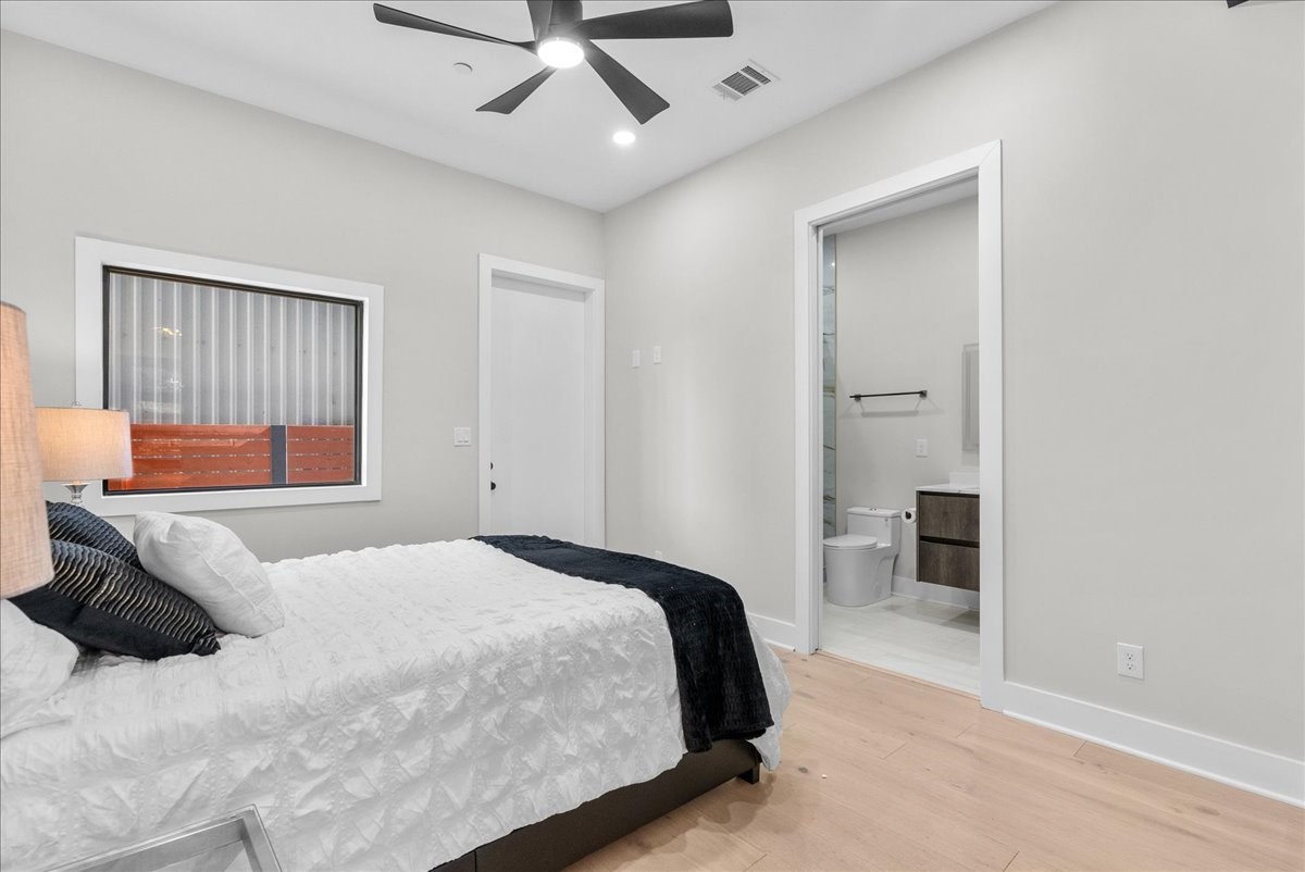 516 Reinerman Street Houston, TX 77007 - Photo 38 of 42 This is a modern bedroom with light walls and wood flooring, featuring a cozy bed, a ceiling fan, and a window. It includes an en-suite bathroom with a sleek vanity and toilet, providing convenience and style.
