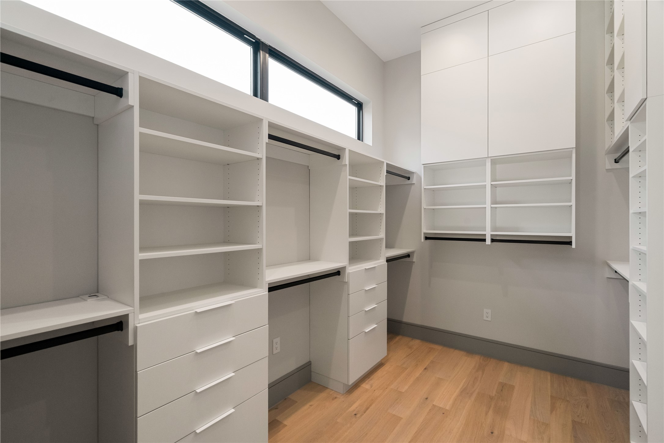 47 Pronghorn Place Spring, TX 77389 - Photo 18 of 44 Spacious walk-in closet with ample shelving and drawer space, featuring sleek white cabinetry and natural wood flooring. High windows provide natural light, enhancing the room's modern and organized feel.