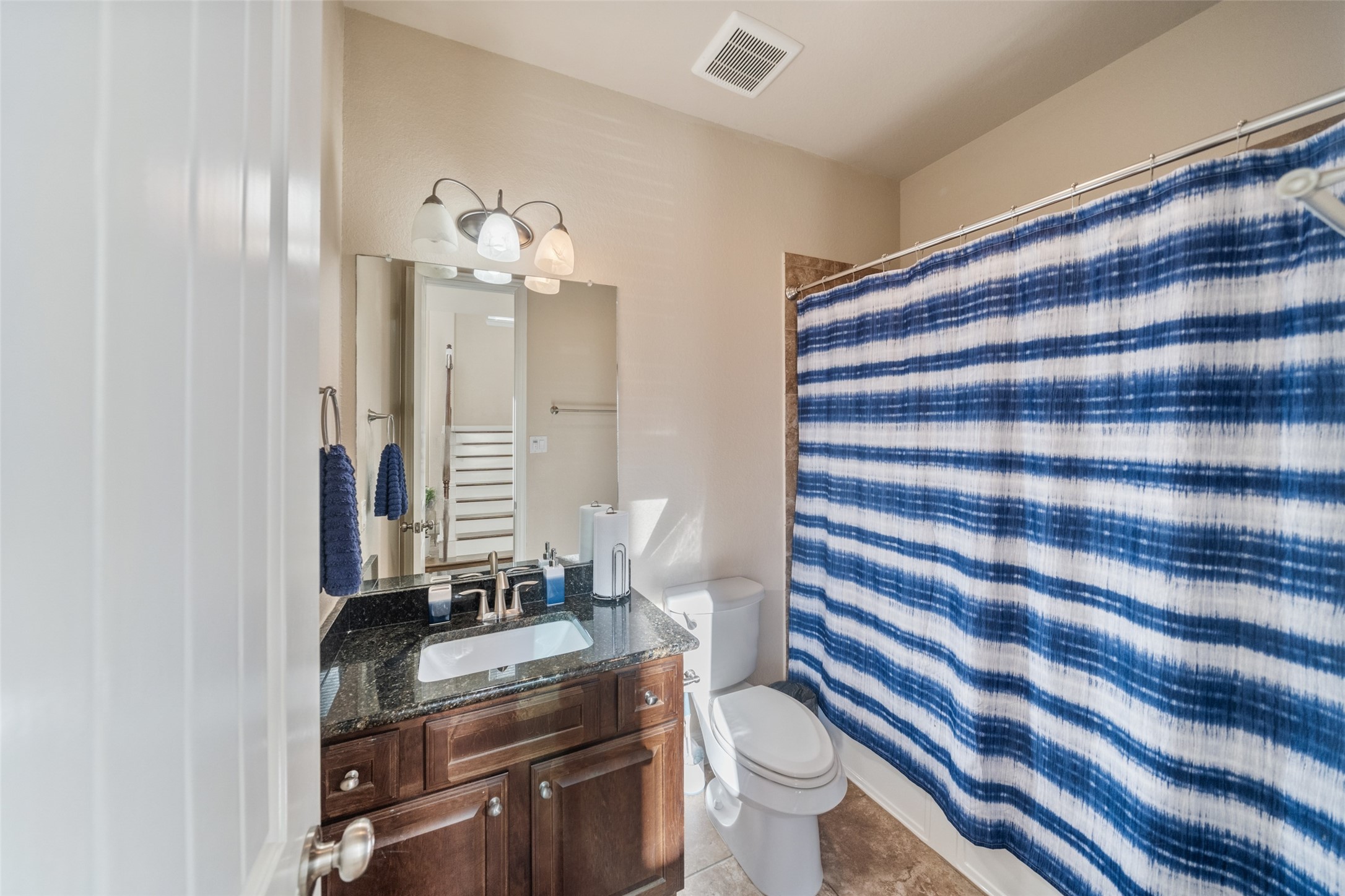 4747 Abercorn Street Rosharon, TX 77583 - Photo 7 of 46 This bathroom features a granite countertop with a modern sink and faucet, complemented by a dark wood vanity. It has a full-sized mirror, overhead lighting, and a shower-tub combo with a blue-striped curtain. The space is well-lit and has neutral wall tones.