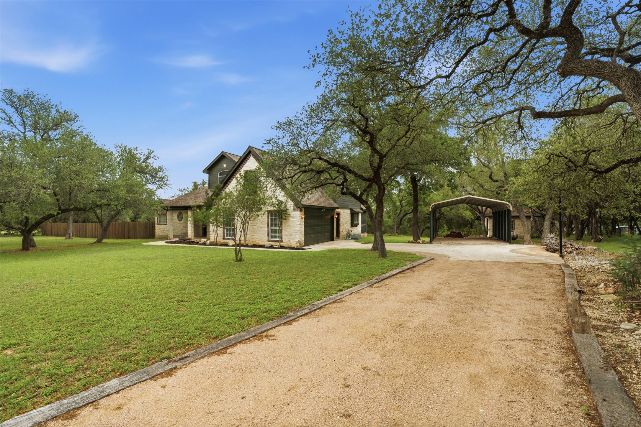 1301 County Road 262 Georgetown, TX 78633 - Photo 3 of 39 Stone exterior residence featuring a gravel driveway, attached carport, mature trees, and a wooden fence