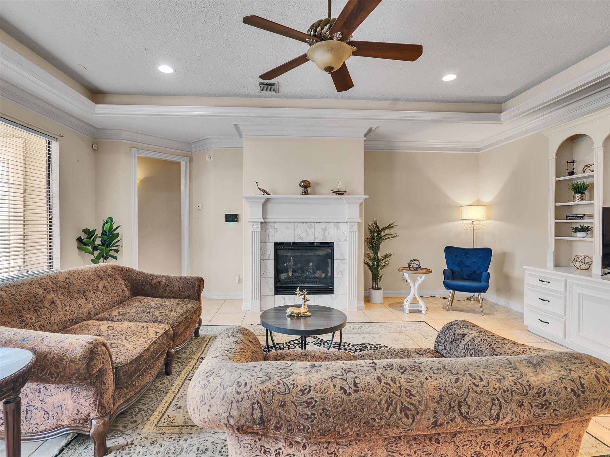 6914 Sandwedge Point Court Spring, TX 77389 - Photo 18 of 50 This inviting living room features a cozy fireplace, elegant crown molding, and built-in shelving. It has a classic feel with patterned sofas, a central coffee table, and a stylish blue accent chair. Natural light flows in through large windows, enhancing the warm, neutral tones.