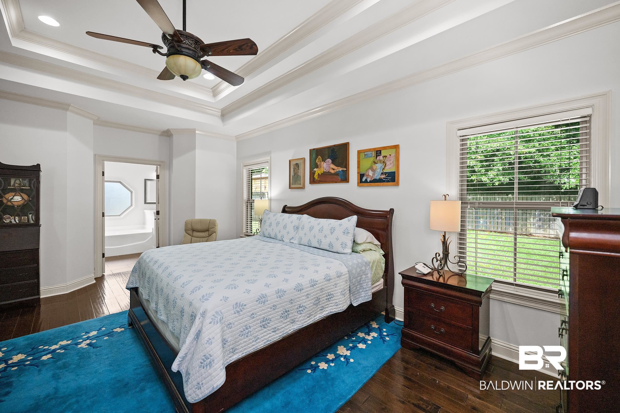 313 Pecan Ridge Boulevard Fairhope, AL 36532 - Photo 12 of 51 Bedroom featuring dark wood-finish flooring, a tray ceiling with a ceiling fan, and a window with blinds overlooking greenery