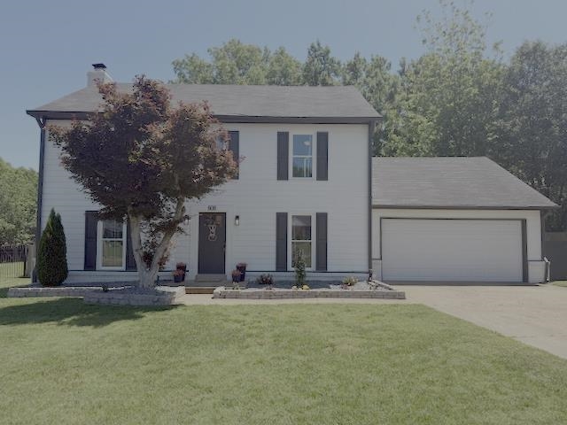 Two-story residence featuring white Hardie board siding, black window shutters, a two-car garage with an asphalt driveway, mature front yard landscaping with a stone retaining wall, and a black front door with decorative lighting
