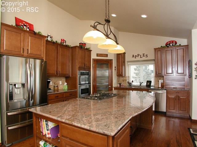 9863 Rockingham Drive Peyton, CO 80831 - Photo 6 of 21 Beautiful gourmet kitchen with vaulted ceiling, slab granite counters with custom tile backsplash, oversized island, 42” maple cabinets with cherry stain & crown molding, crumb catcher, 2 built-in spice racks, walk-in pantry with glass door & appliance garage.