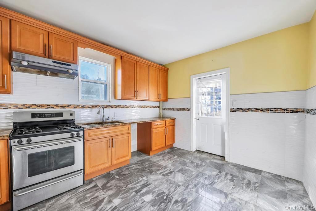 95-26 86th Street Queens, NY 11416 - Photo 8 of 28 Kitchen featuring stainless steel gas range, under cabinet range hood, brown cabinets, light stone countertops, and a wainscoted wall
