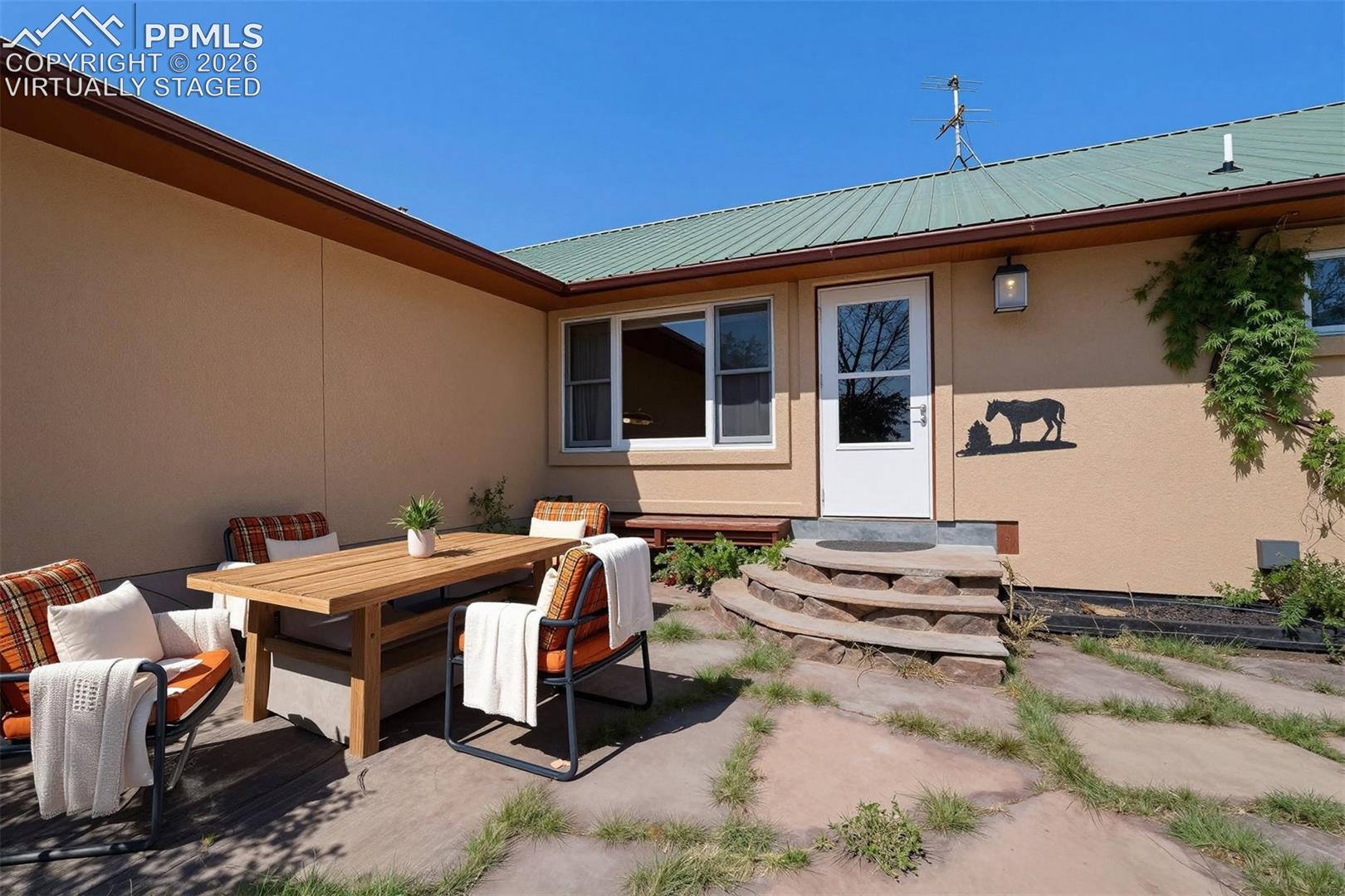 2550 Slocum Road Peyton, CO 80831 - Photo 41 of 50 virtually staged Outdoor patio area featuring large flagstone pavers, a light tan stucco exterior, a green metal roof, and a white entry door with an exterior light fixture