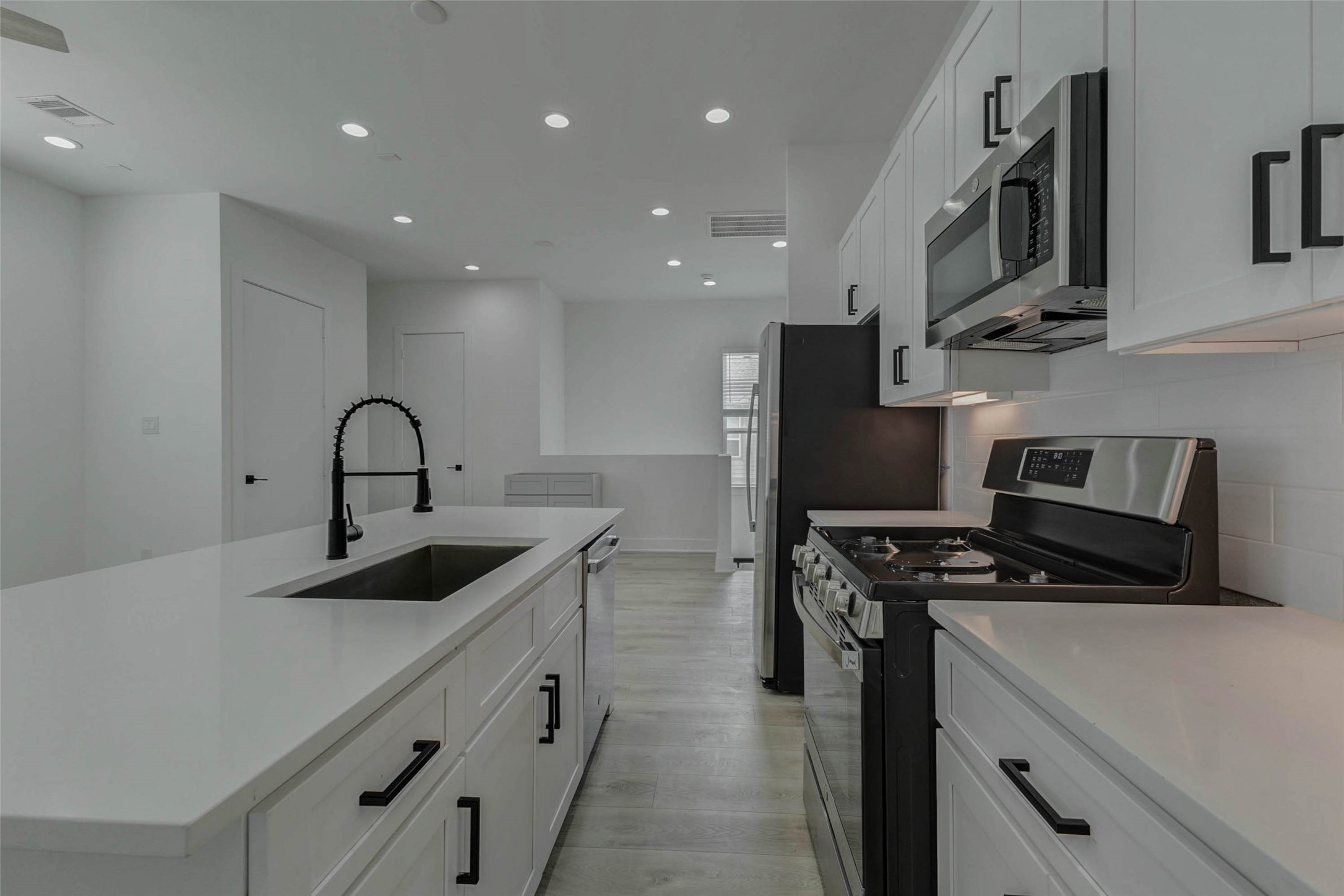 5705 Elysium Street Houston, TX 77091 - Photo 15 of 22 Kitchen close-up featuring a large island sink, matte black pull-down faucet, stainless gas range, built-in microwave, refrigerator, white shaker-style cabinetry, black hardware, and bright under-cabinet lighting. The clean finishes and open sightline create a crisp, functional, modern cooking space.