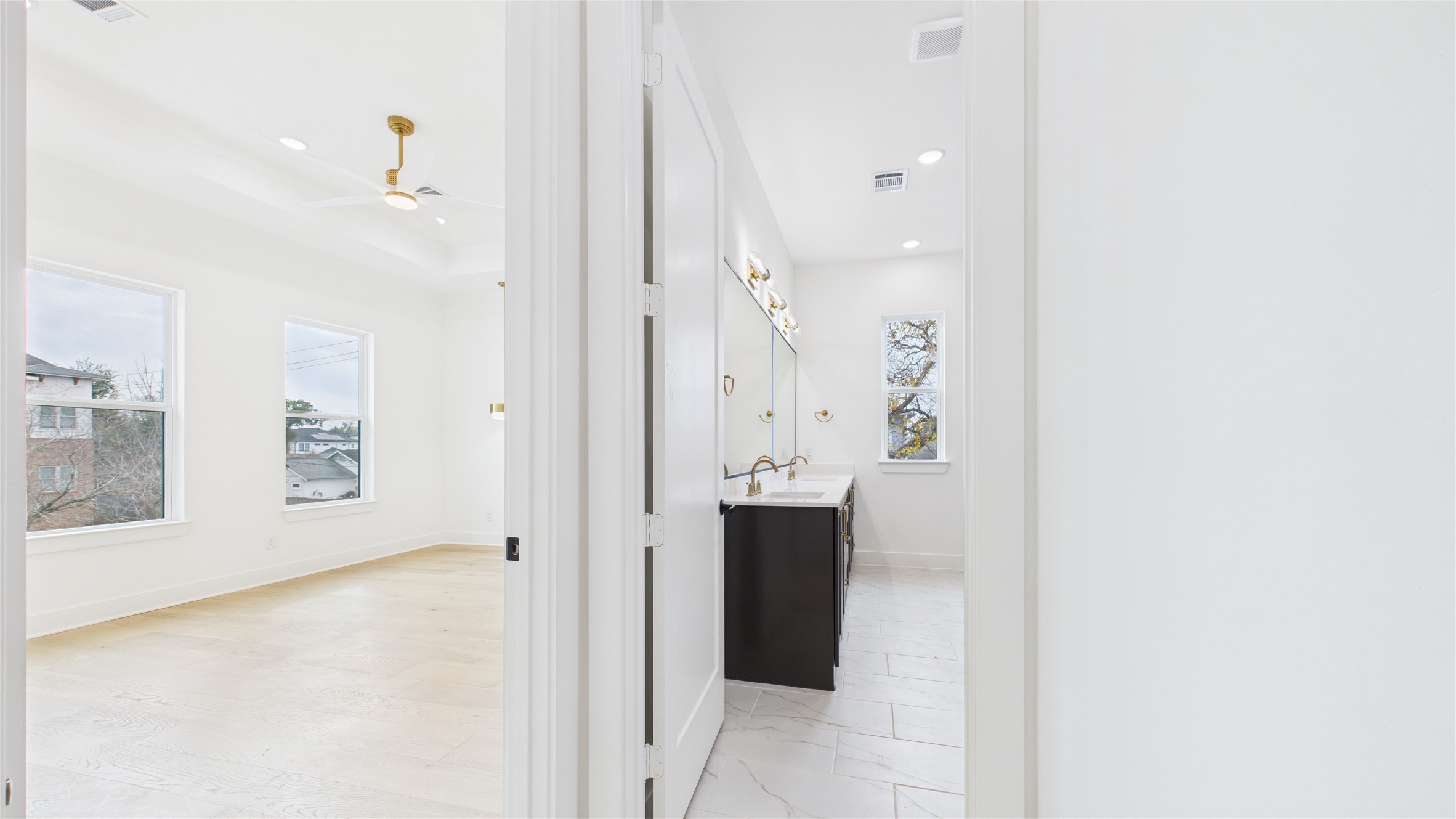 602 Jewett Street, Unit A Houston, TX 77009 - Photo 22 of 46 This photo shows a bright, modern bathroom connected to a spacious bedroom. The bathroom features a sleek double vanity, large mirrors, and ample natural light, while the bedroom boasts large windows and a ceiling fan, creating an airy and inviting space.