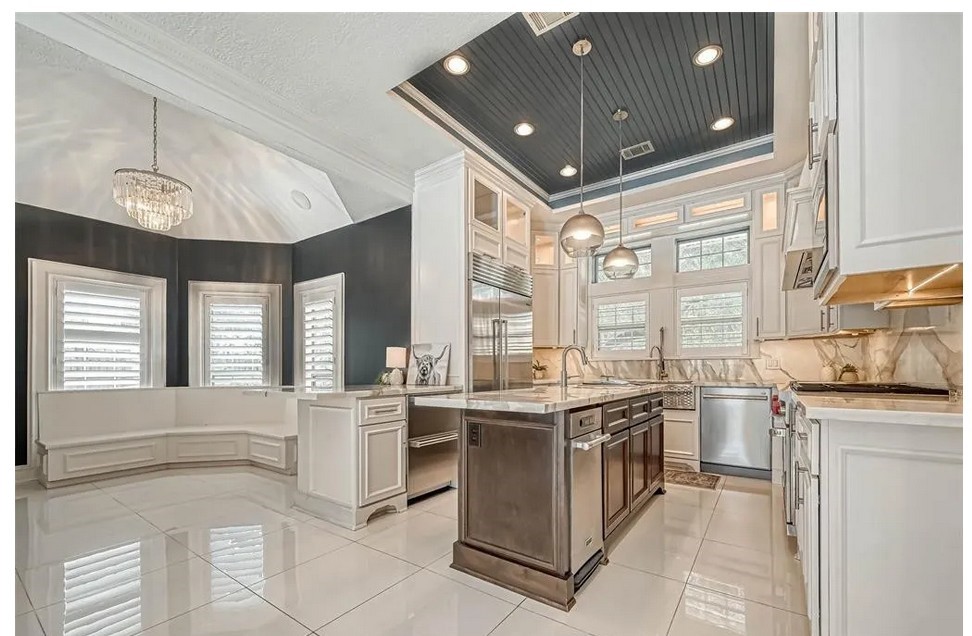 263 Wood Loop Street Houston, TX 77015 - Photo 18 of 45 This kitchen features a modern design with sleek white cabinetry, a large central island, and stainless steel appliances. The high ceiling and ample windows provide abundant natural light, complemented by elegant pendant lights and a chandelier. The space includes a cozy breakfast nook with bay windows, offering a stylish and functional setting.