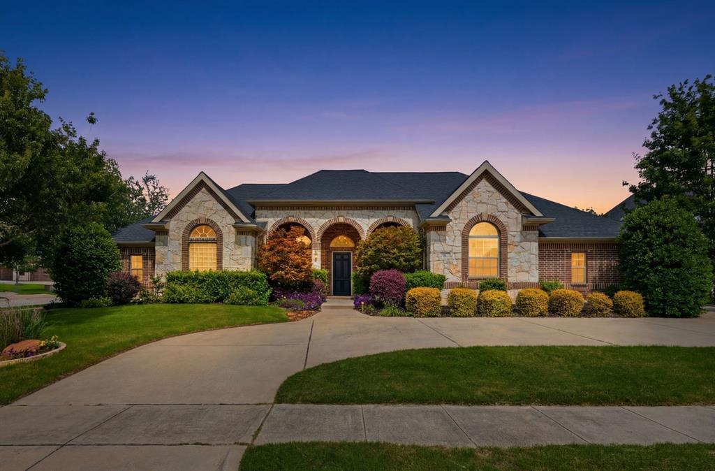 7013 Handel Colleyville, TX 76034 - Photo 1 of 37 Stone and brick exterior featuring arched windows, a dark shingle roof, mature landscaping, and an expansive concrete driveway