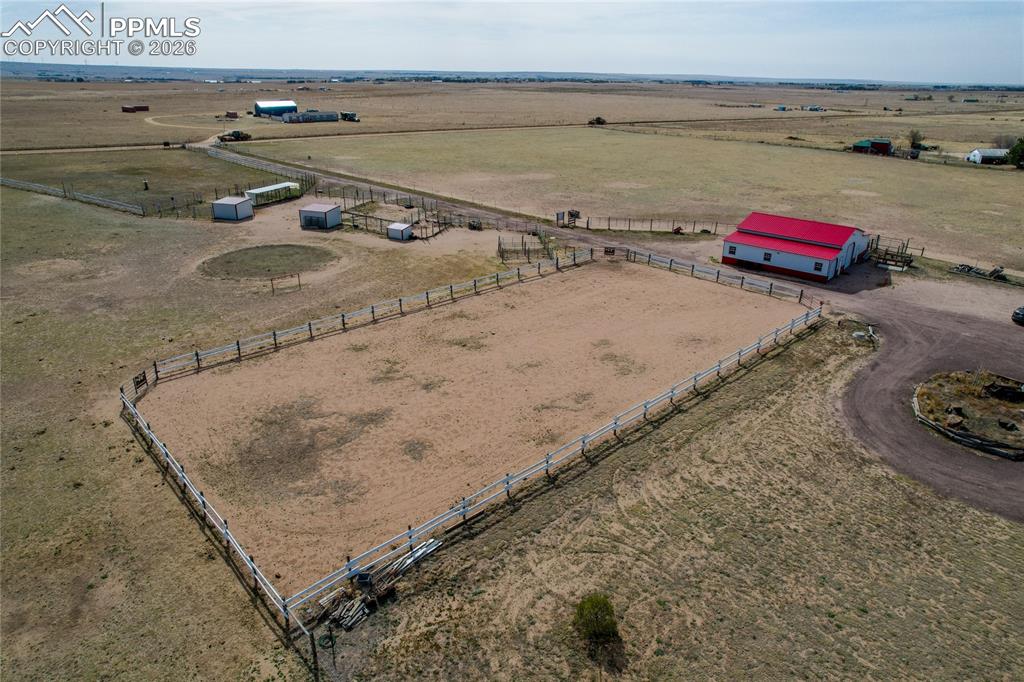 7880 J D Johnson Road Peyton, CO 80831 - Photo 34 of 50 Expansive outdoor arena featuring perimeter fencing, an adjacent barn with a red roof, a circular driveway, and additional outbuildings