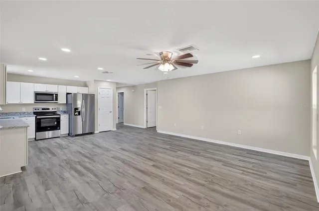 an empty room with wooden floor kitchen view and a ceiling fan