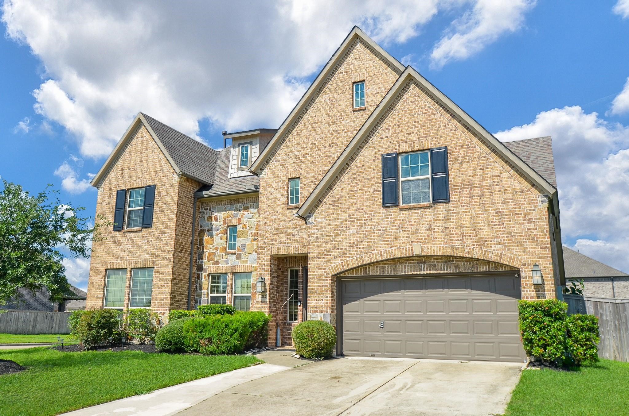 This is a two-story brick home with a pitched roof and a two-car garage. It features a mix of large and small windows with shutters, a well-maintained front yard with shrubs, and a concrete driveway. The facade combines brickwork with stone accents, creating a classic and inviting appearance.