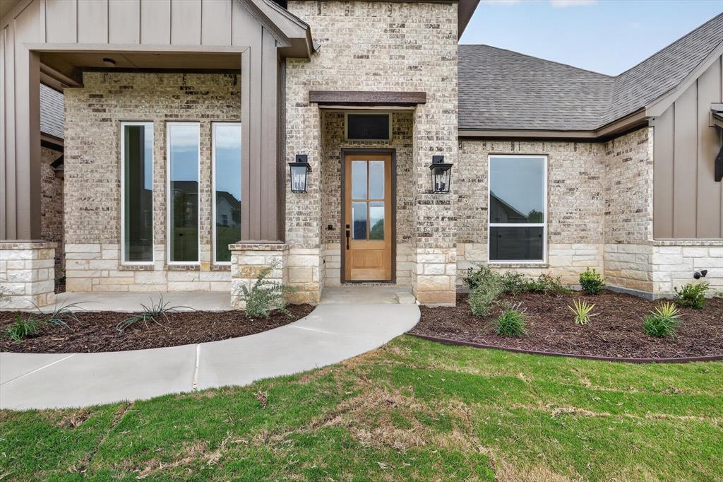 471 Red Bird Drive Springtown, TX 76082 - Photo 3 of 35 Brick and stone exterior featuring vertical panel siding accents, an inviting entryway with a wood-finish door, exterior wall lanterns, and a concrete pathway