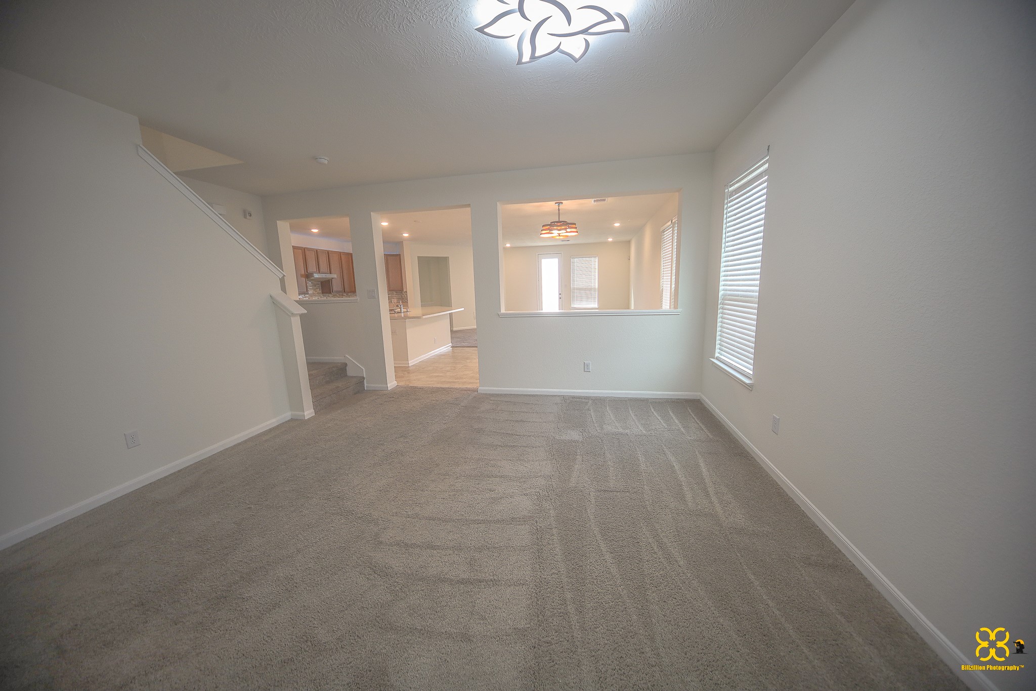 5820 Bluebonnet Lane Rosenberg, TX 77469 - Photo 12 of 43 This photo shows a spacious, open-plan living area with carpeted floors and neutral walls. It features a view into a kitchen with a breakfast bar, plenty of natural light from large windows, and an elegant ceiling light fixture.