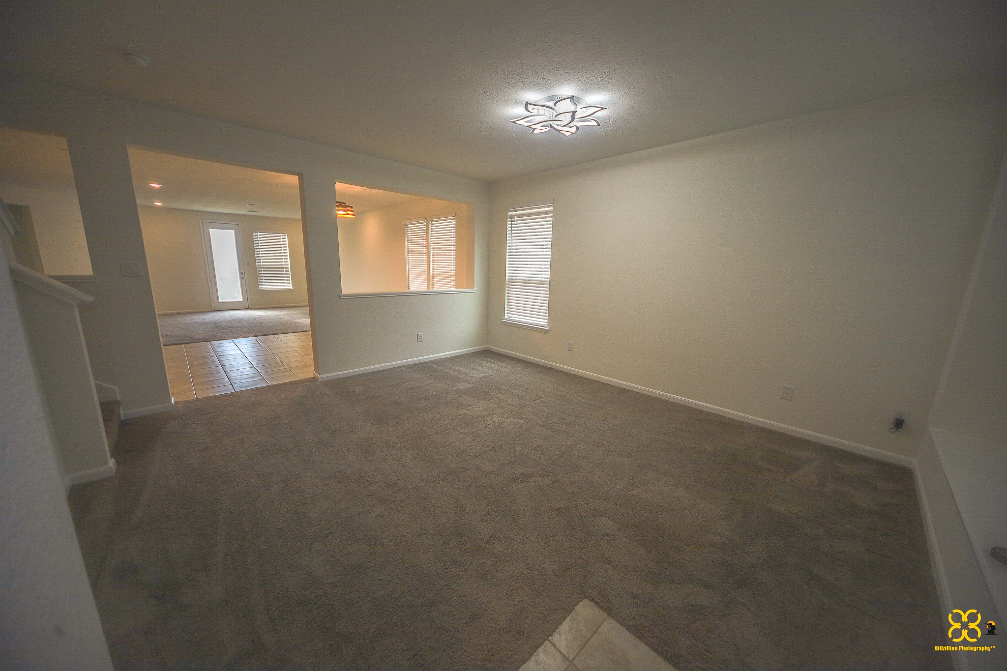 5820 Bluebonnet Lane Rosenberg, TX 77469 - Photo 13 of 43 This photo shows a spacious living area with carpeted flooring, neutral walls, and a modern light fixture. Large windows allow natural light, and there's an open layout leading to a tiled section, possibly a dining area, with access to the outdoors through glass doors.