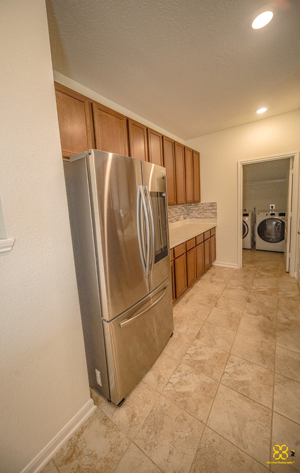 5820 Bluebonnet Lane Rosenberg, TX 77469 - Photo 20 of 43 This photo shows a modern kitchen area with stainless steel appliances, including a refrigerator. The kitchen features ample wooden cabinetry and a tiled floor. Beyond the kitchen, there's a view into a laundry room with a washer and dryer.