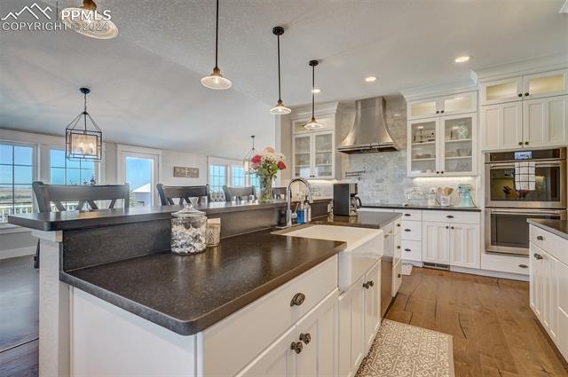 24076 Kobilan Road Calhan, CO 80808 - Photo 11 of 50 Kitchen with light hardwood / wood-style flooring, white cabinets, double oven, an island with sink, and wall chimney exhaust hood