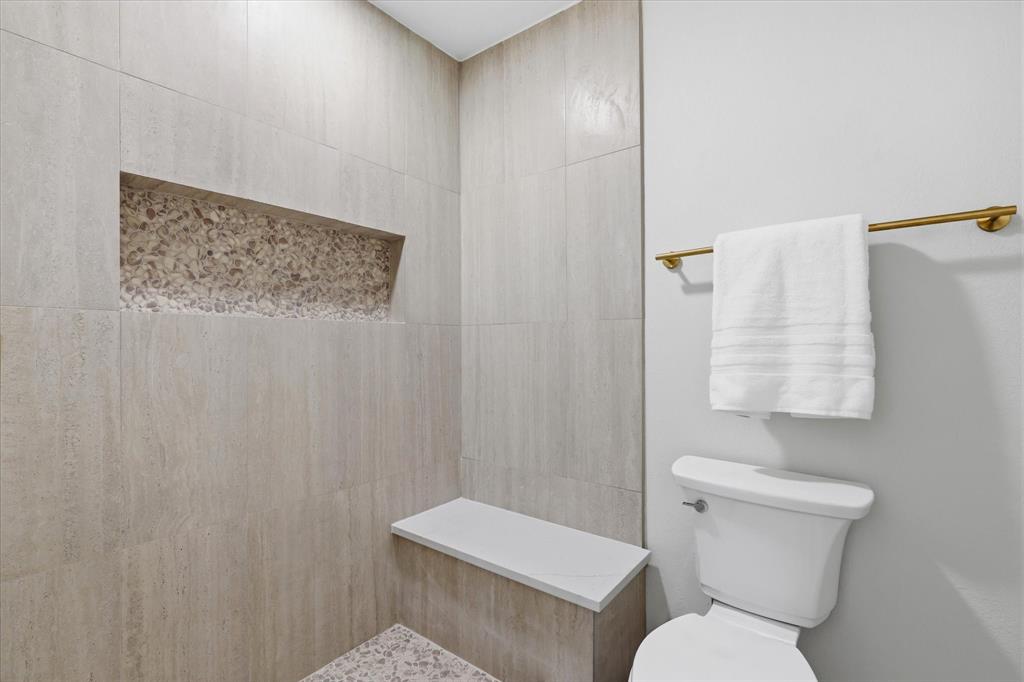 3405 Mill Ridge Street Bedford, TX 76021 - Photo 19 of 32 Tiled shower with a built-in niche featuring decorative stone, an integrated shower bench with a solid surface top, and a white toilet with a brass towel bar