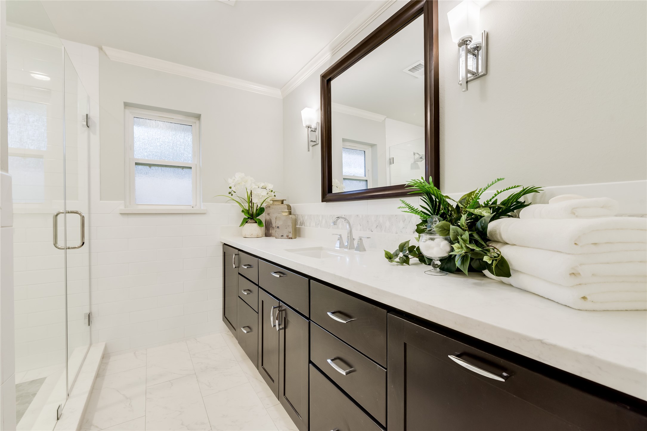 1010 Thornton Road Houston, TX 77018 - Photo 21 of 27 This modern primary bathroom features dark wood cabinets,quartz counter tops and a large framed mirror. It has a spacious glass-enclosed shower,and stylish fixtures. Natural light filters through frosted windows, enhancing the bright and fresh ambiance.