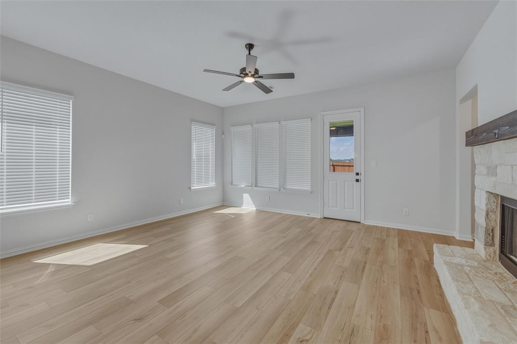 1213 Colca Drive Springtown, TX 76082 - Photo 9 of 23 Spacious living area featuring light wood-finish flooring, a stone-clad fireplace with a wood mantle, multiple windows with blinds, a ceiling fan with integrated lighting, and a rear exit door