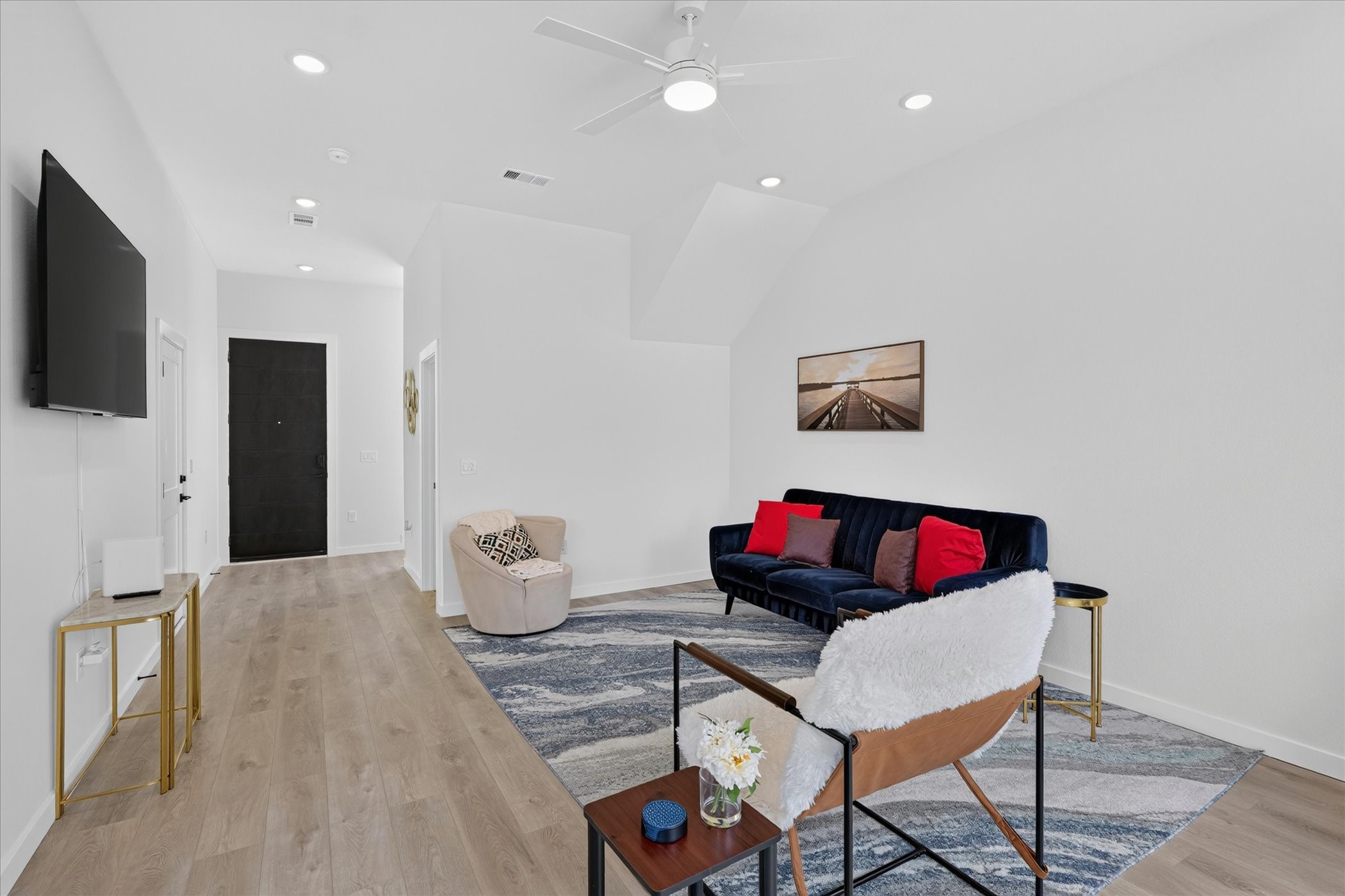 Bright and modern living area with high ceilings, a cozy seating arrangement, and stylish decor. Features a wall-mounted TV, wood flooring, and a neutral color palette, creating an inviting atmosphere.