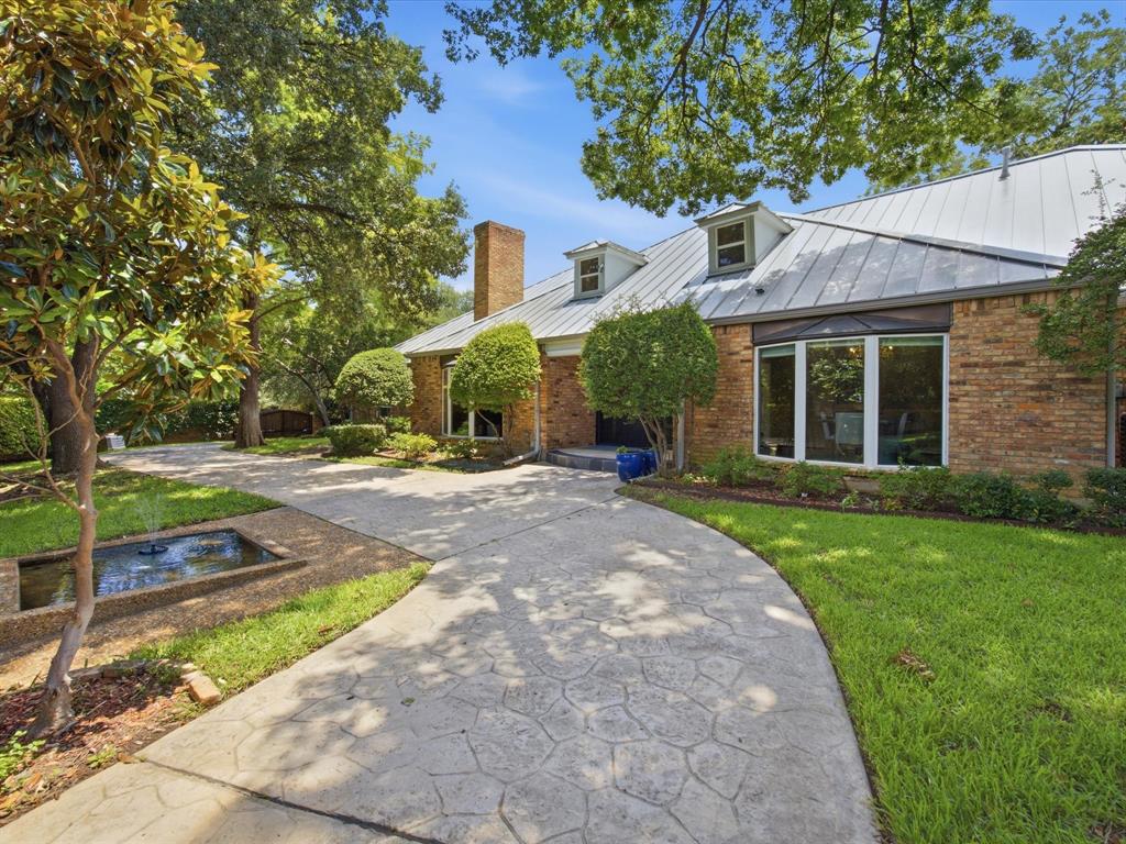6160 Spring Valley Road Dallas, TX 75240 - Photo 2 of 40 Brick exterior featuring a metal roof with dormer windows, an imprinted concrete driveway, mature trees, and a decorative fountain