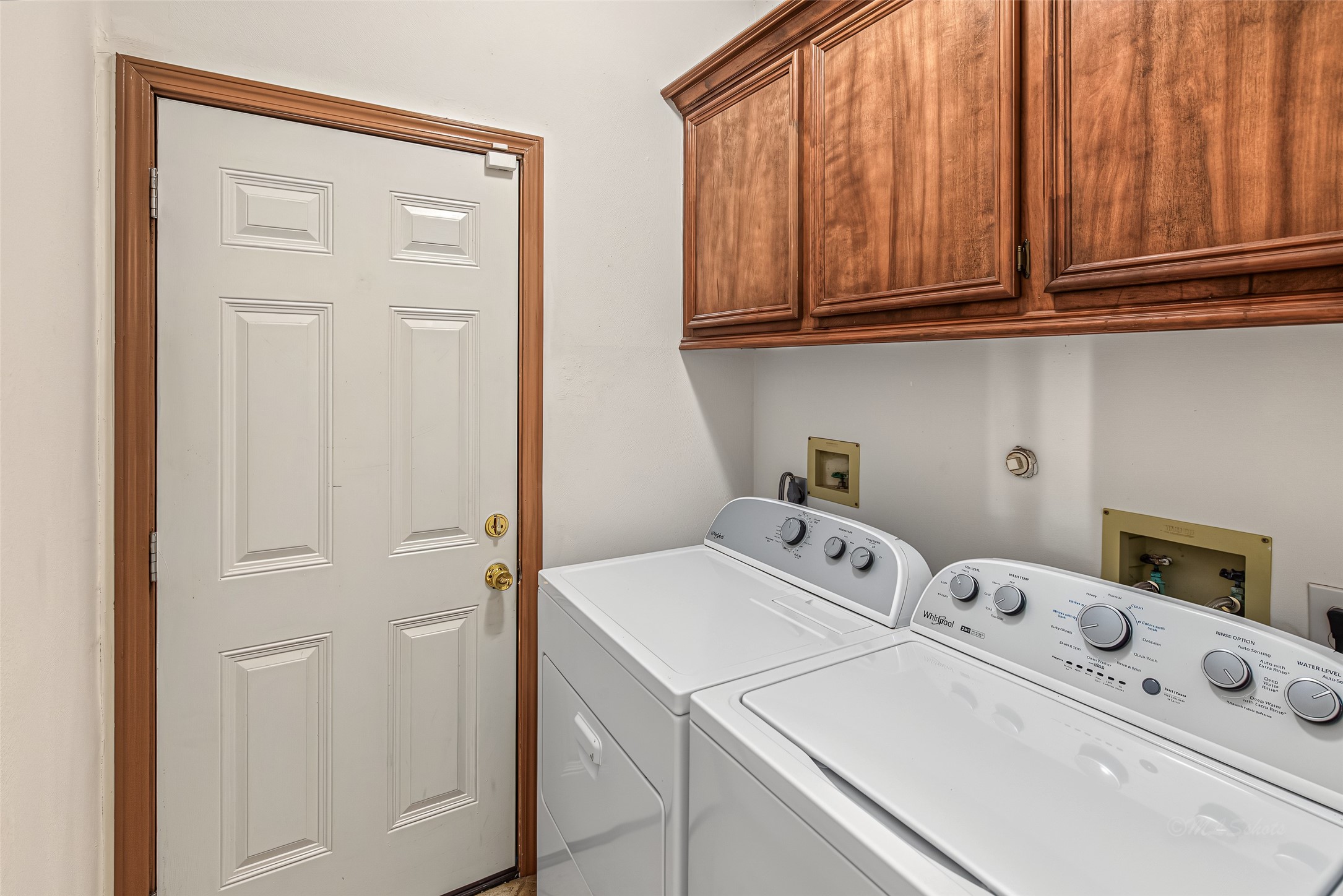 1345 Peach Ridge Road Brookshire, TX 77423 - Photo 14 of 50 Designed for maximum efficiency, this functional utility room features rich, custom-finished upper wood cabinetry that provides ample concealed storage for laundry supplies and everyday essentials. Situated directly alongside a white top-loading washer and dryer, the space is neatly finished with matching wood casing around the interior door, blending seamlessly with the home's cohesive millwork design.