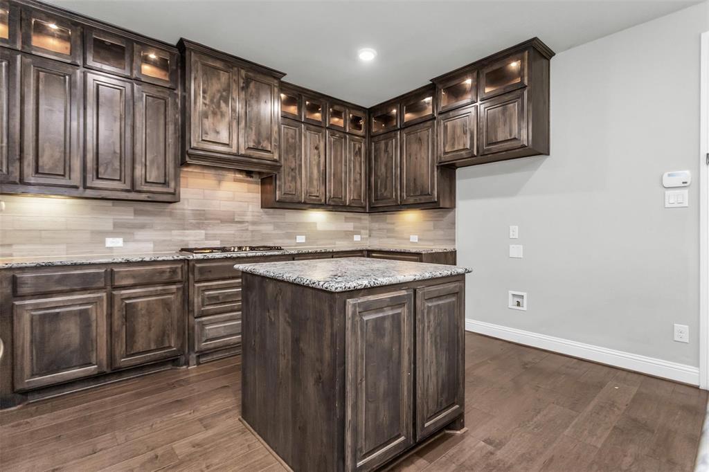 6333 Hall Road Frisco, TX 75034 - Photo 16 of 38 Wood-finish flooring, dark wood cabinetry with integrated lighting, a central island with a stone countertop, a tile backsplash, and built-in appliances